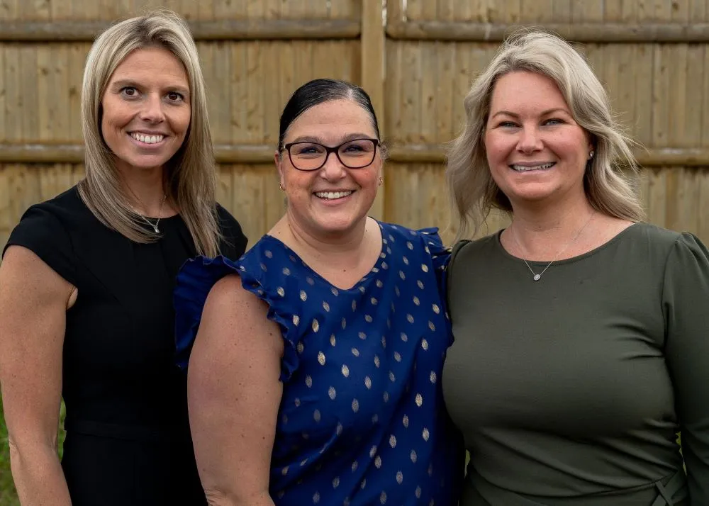 Three smiling women standing together in front of a wooden fence